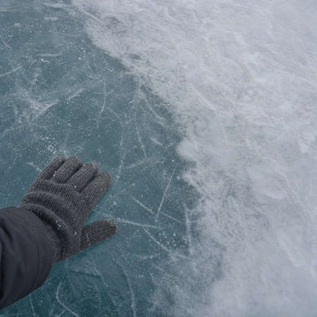 Close-up of a frozen lake surface showing clear bluish ice next to cloudy white opaque patches, with a gloved hand for scale